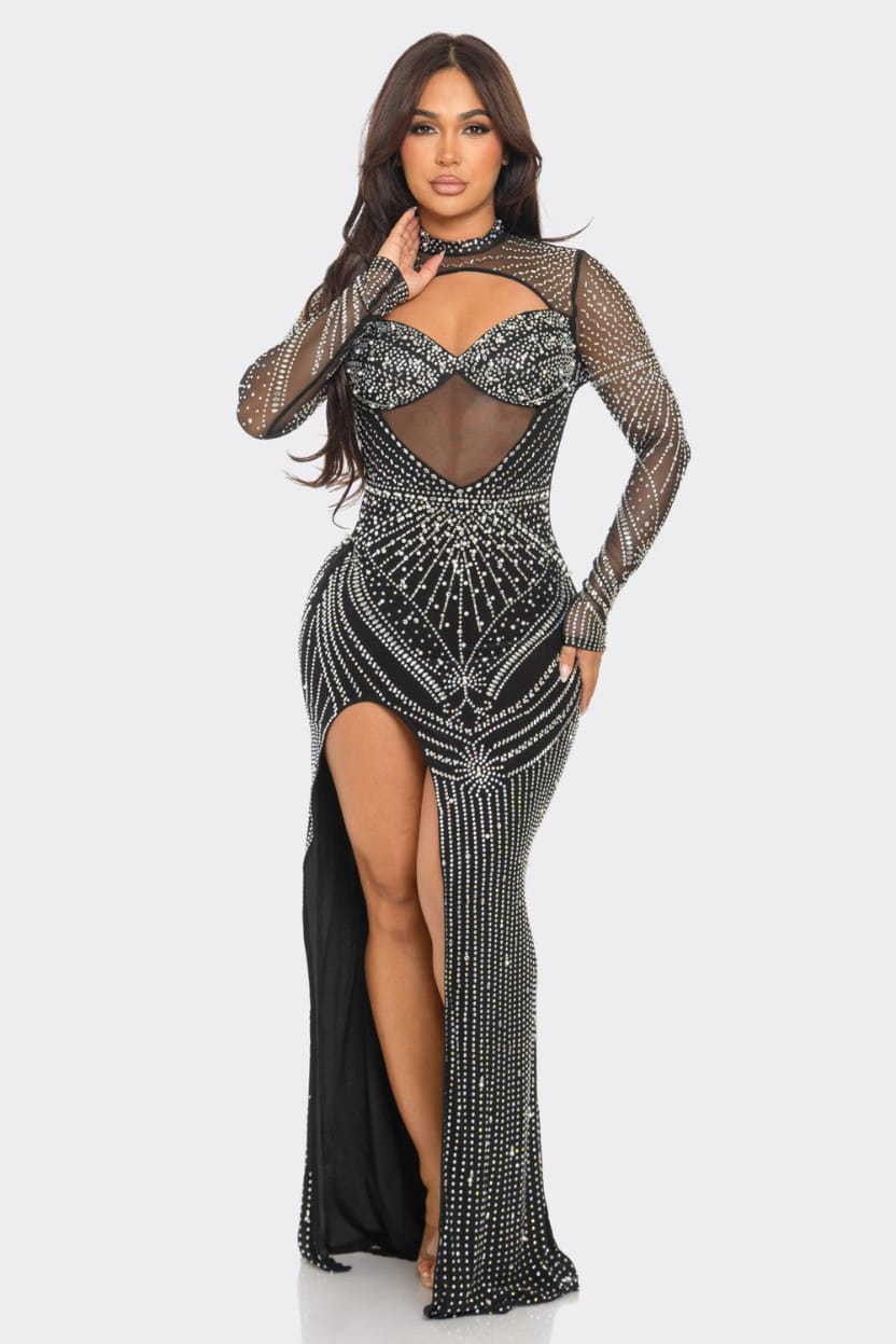 A woman modeling a long-sleeve, floor-length black gown covered in silver rhinestone embellishments, featuring a sheer mesh bodice with a sweetheart cutout and a high thigh slit, against a light gray studio background.