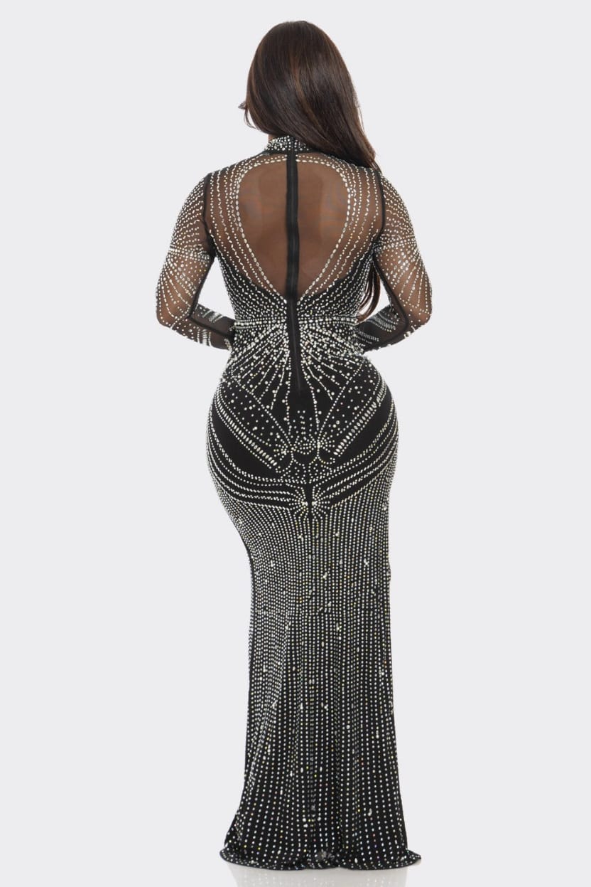A woman viewed from the back wearing a fitted black evening gown covered in silver rhinestone embellishments, with sheer mesh sleeves and a high open back, against a light gray studio background.