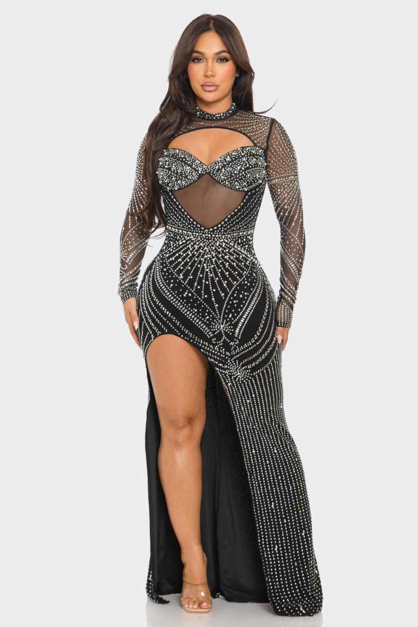 A plus-size model wearing a long-sleeved black maxi gown covered in silver rhinestone and sequin embellishments, featuring a sheer mesh bodice with a sweetheart cutout and a high leg slit, set against a plain light gray background.