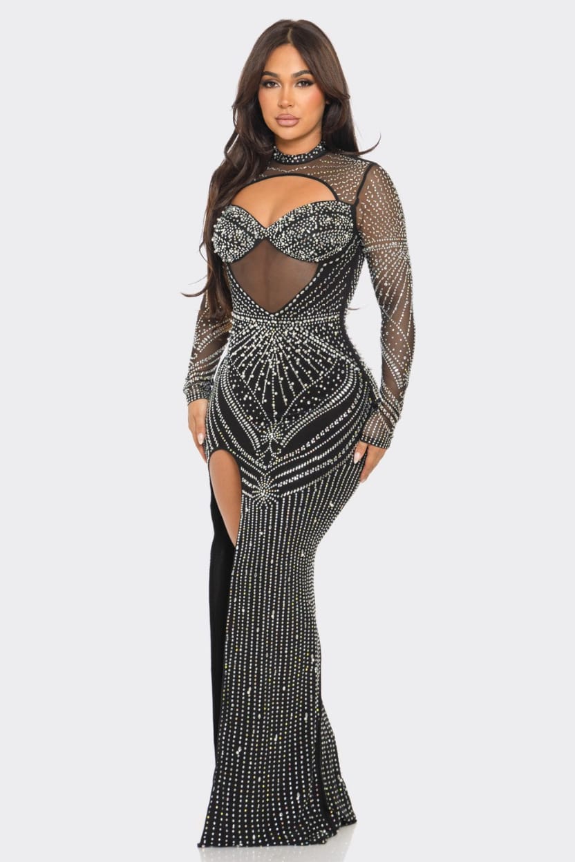 A woman modeling a long-sleeve, floor-length black gown covered in silver rhinestone embellishments, featuring a sheer mesh bodice with a sweetheart cutout and a high thigh slit, against a light gray background.