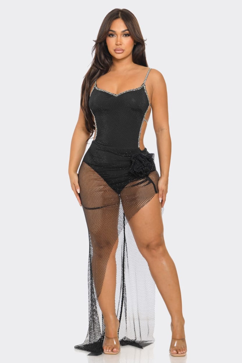A woman modeling a black lingerie bodysuit with sheer fishnet panels and a long trailing net skirt, photographed against a light gray studio background.