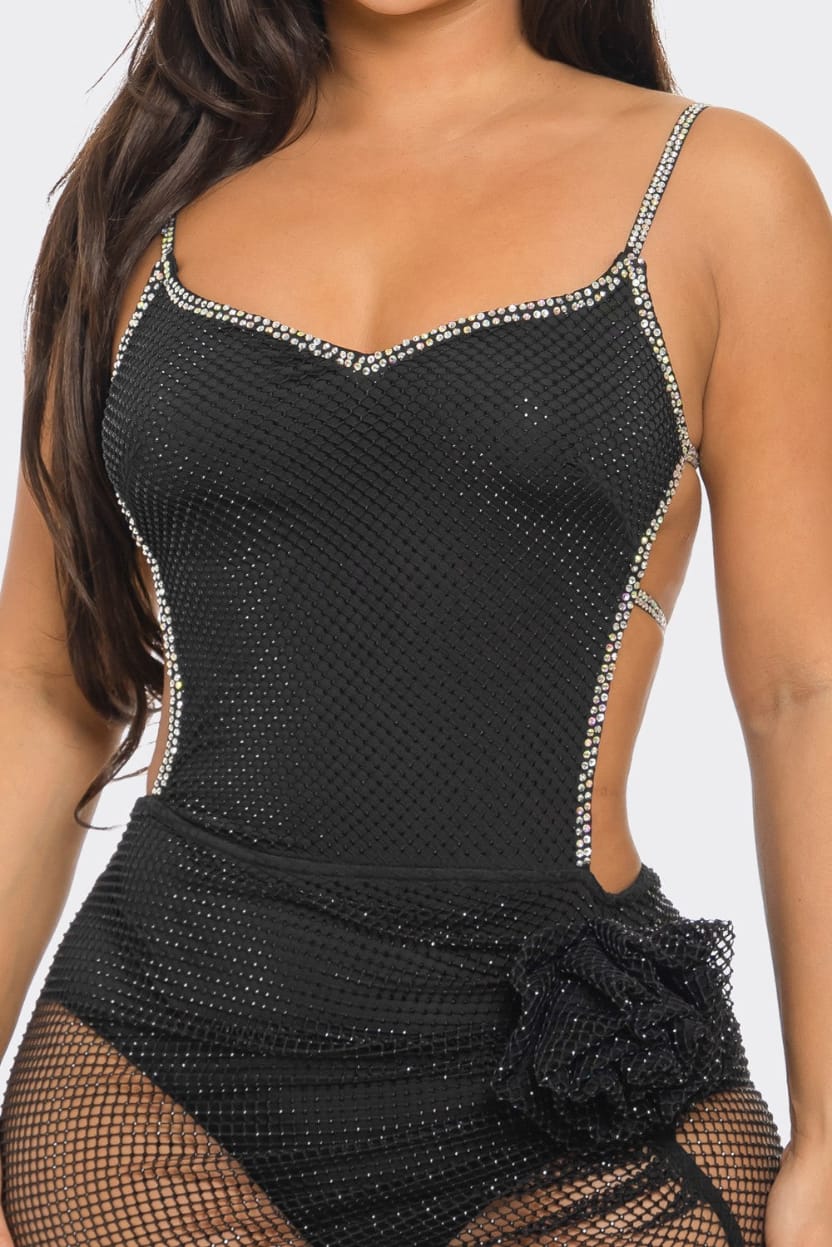 A close-up of a woman wearing a black mesh bodysuit with silver rhinestone trim along the straps and neckline, plus a matching black fishnet skirt with a ruffled overlay.