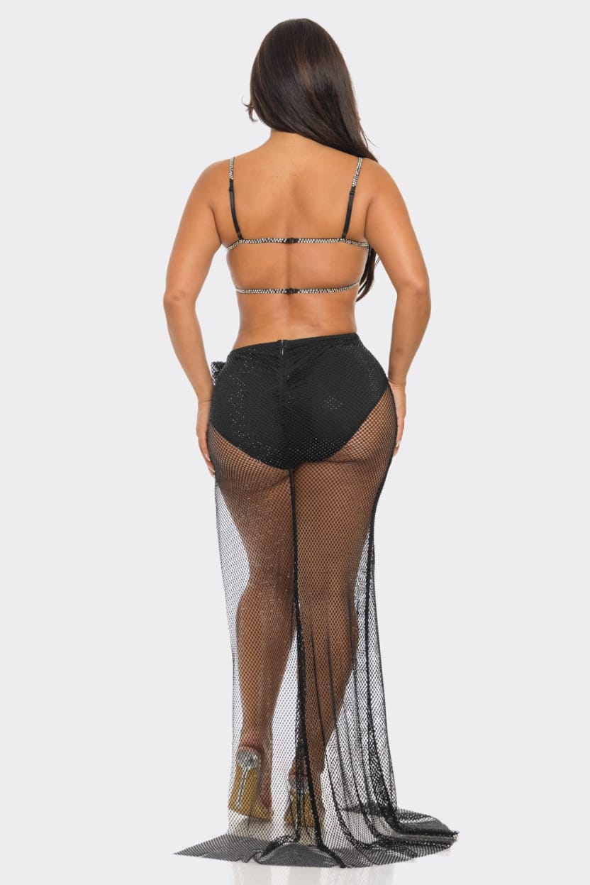 A woman shown from the back wearing a black strappy bikini-style top with rhinestone trim, black high-waisted shorts, sheer black fishnet stockings, and a long black mesh train with rhinestone embellishments, against a light gray studio background.