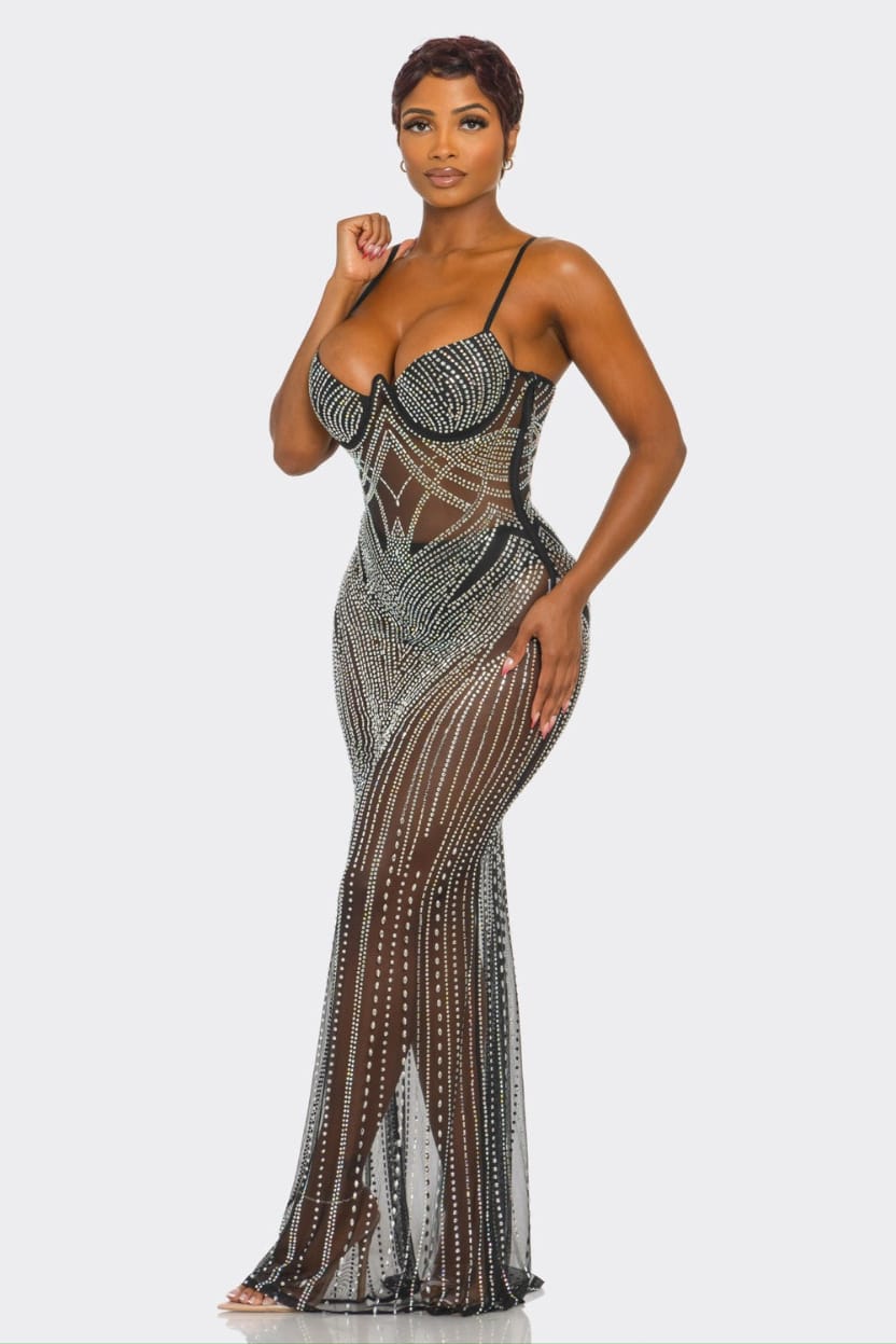 A woman modeling a sheer black and silver beaded maxi dress with a structured corset-style bodice and flared skirt, photographed against a light gray studio background.