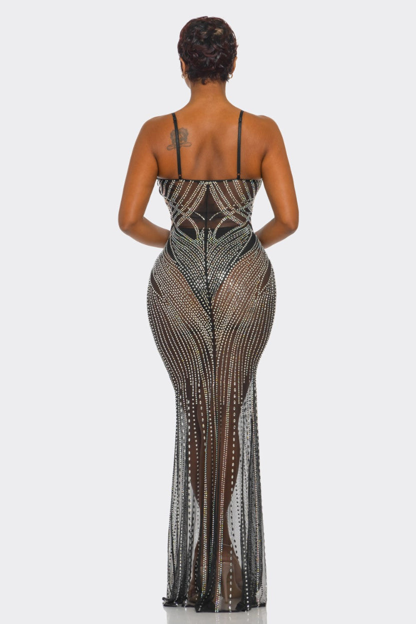 A woman viewed from the back wearing a sheer black mesh maxi dress heavily embellished with silver rhinestones, set against a plain light gray studio background.
