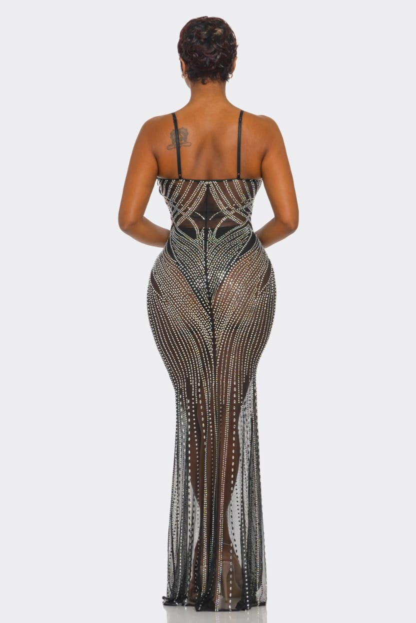 A woman viewed from the back wearing a sheer black mesh maxi dress heavily embellished with silver rhinestones, set against a plain light gray studio background.