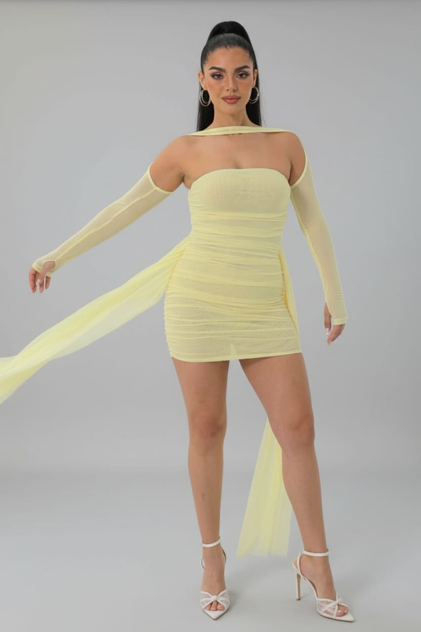 A woman modeling a pale yellow off-the-shoulder mini dress with long sheer sleeves and a flowing sheer train, paired with white strappy high-heeled sandals in a bright white room.