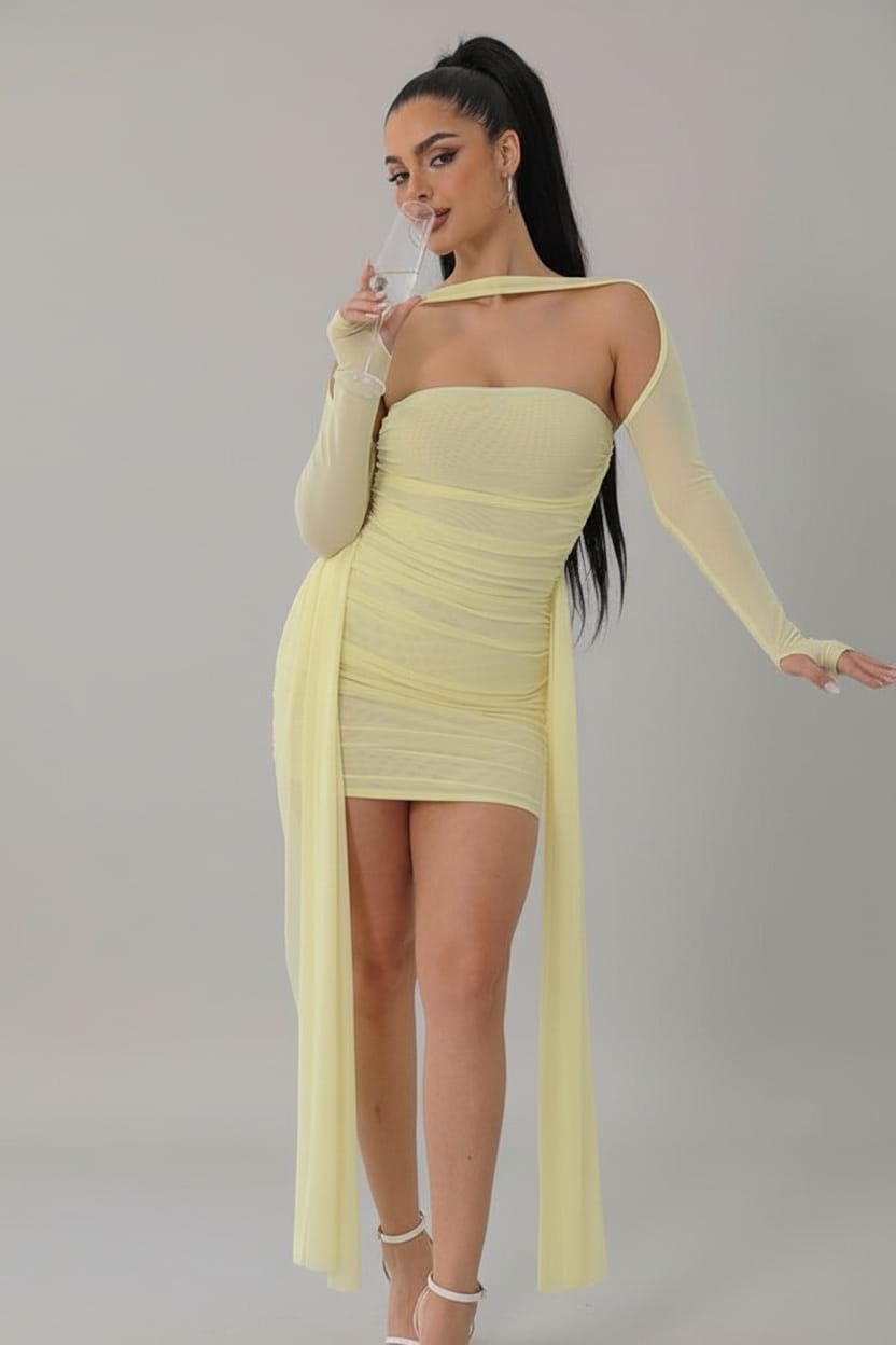 A woman models a pale yellow strapless mini dress with long sheer sleeves and a draped overlay, standing in a bright white room next to a gold-framed mirror.