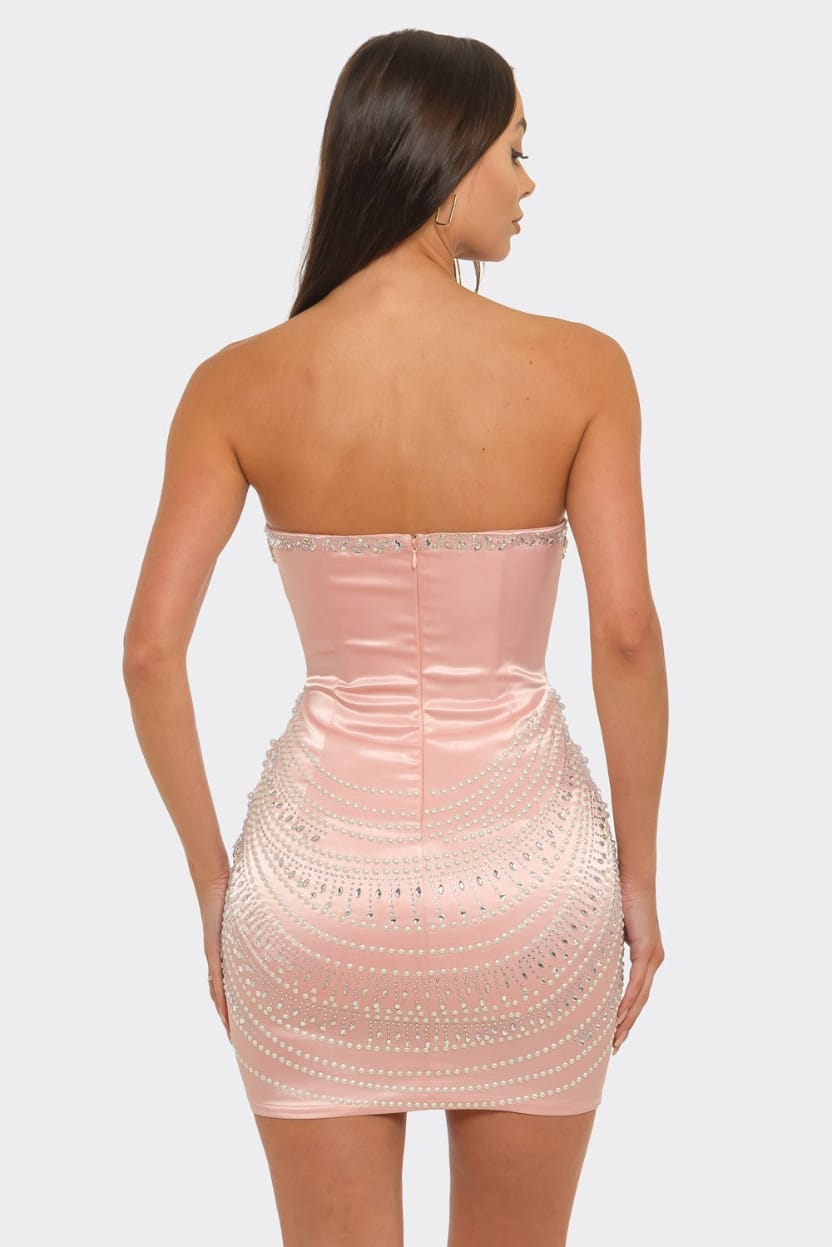 A woman shown from the back wearing a strapless blush pink bodycon mini dress with rhinestone embellishments, set against a plain light gray studio background.