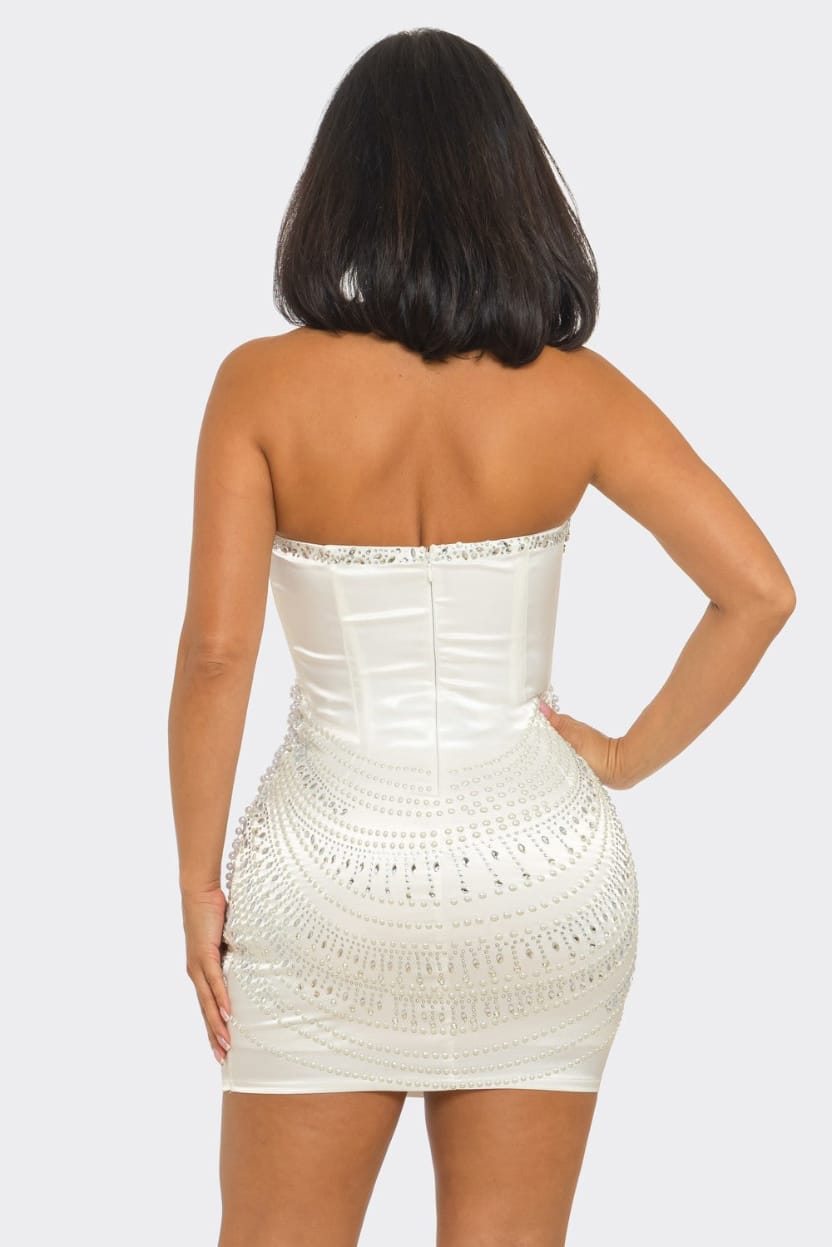 A woman shown from the back wearing a strapless white mini dress with a structured corset-style back and embellished beaded details, against a light gray studio background.