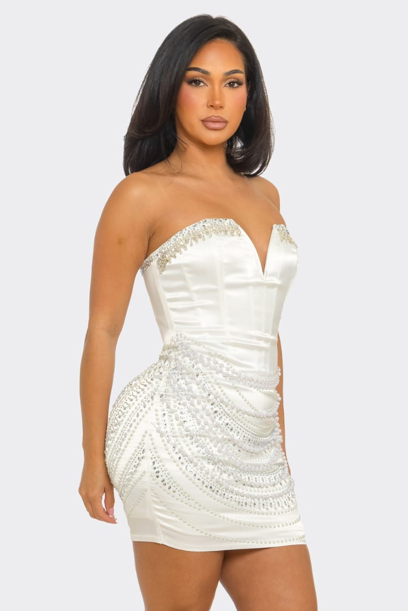A woman modeling a strapless white mini dress with a sweetheart neckline, structured corset-style bodice, and cascading beaded embellishments on the skirt, against a light gray studio background.