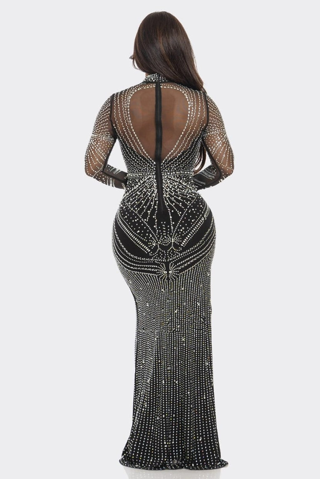 A woman viewed from the back wearing a fitted black evening gown covered in silver rhinestone embellishments, with sheer mesh sleeves and a high open back, against a light gray studio background.