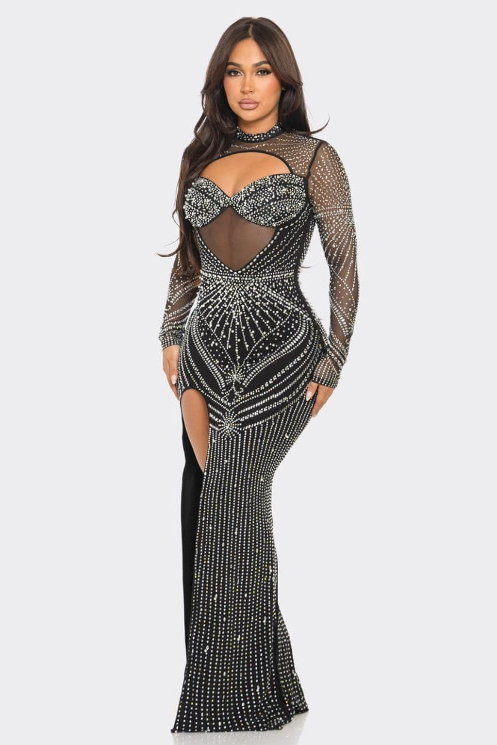 A woman modeling a long-sleeve, floor-length black gown covered in silver rhinestone embellishments, featuring a sheer mesh bodice with a sweetheart cutout and a high thigh slit, against a light gray background.