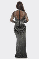 A woman viewed from the back wearing a fitted black evening gown covered in silver rhinestone embellishments, with sheer mesh sleeves and a high open back, against a light gray studio background.