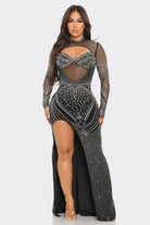 A plus-size model wearing a long-sleeved black maxi gown covered in silver rhinestone and sequin embellishments, featuring a sheer mesh bodice with a sweetheart cutout and a high leg slit, set against a plain light gray background.
