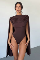 Woman wearing a brown one-piece swimsuit with cape-like design against a plain background