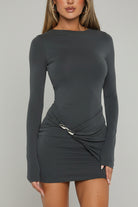 A woman wearing a dark gray long-sleeve bodycon mini dress with a twisted detail at the waist, standing in front of a white door with green garden foliage visible on the right.