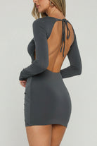 A woman wearing a fitted long-sleeve mini dress with an open back and tie details, shown from the back in a bright indoor setting with a window and greenery outside.