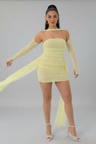 A woman modeling a pale yellow off-the-shoulder mini dress with long sheer sleeves and a flowing sheer train, paired with white strappy high-heeled sandals in a bright white room.
