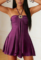 A woman wearing a purple halter mini dress with a ruched bodice, flared skirt, and a large jeweled circular pendant at the neckline.