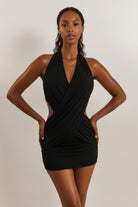 A woman modeling a black halter-neck mini dress with a deep V-neckline, crossover wrap detail at the bust, side cutouts, and gathered draping at the skirt, shown against a plain light beige studio background.