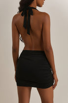 A woman wearing a black halter-neck mini dress with a wrap-style crossover bodice, side cutouts, and gathered draping details, photographed against a plain light beige studio background.