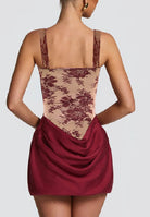 Rear view of a model wearing a short dress with a beige floral lace bodice and a draped burgundy skirt, set against a solid light gray background.