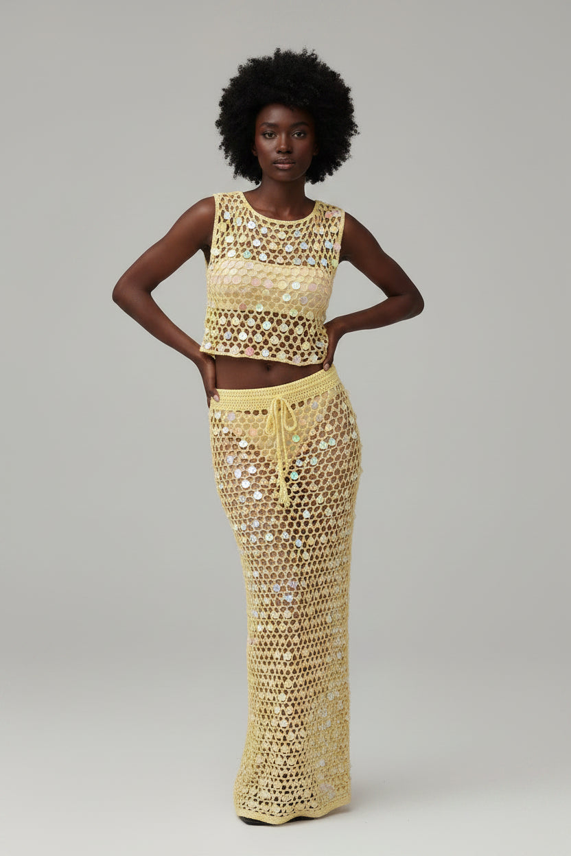Woman wearing a yellow crochet outfit by a palm tree and beach.