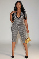 A woman modeling a black and white horizontally striped one-piece jumpsuit with a deep V-neckline and black trim, paired with black pointed-toe heels and a small gold handbag, standing outdoors at night.